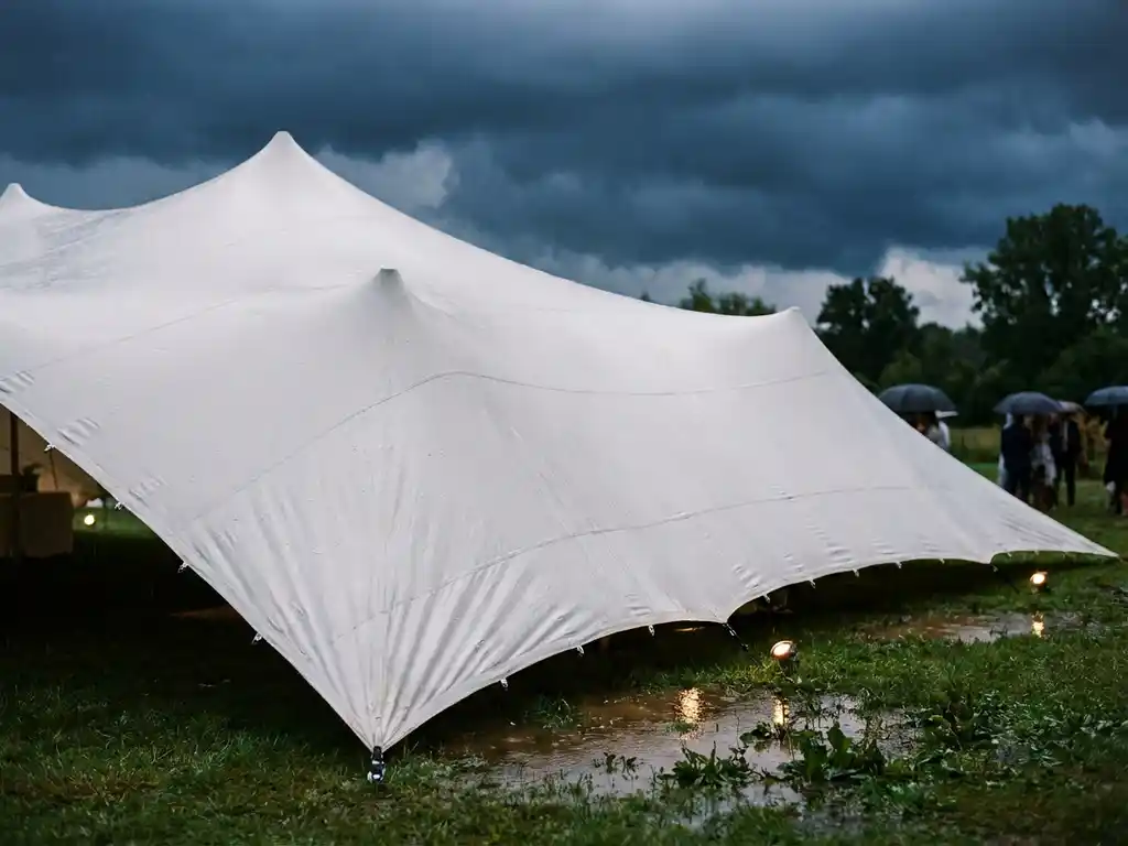 Witte stretchttent met regendruppels op gespannen doek tijdens storm, waterplassen op nat gras onder donkere wolken