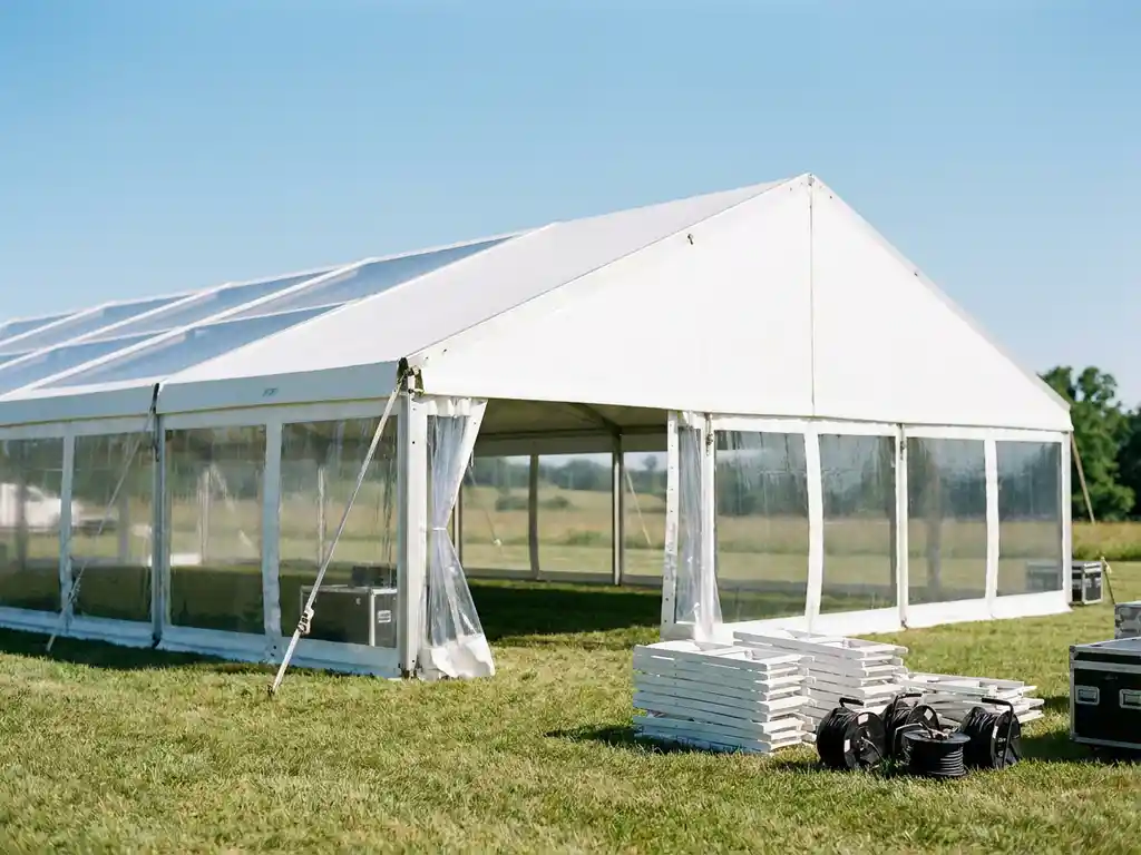 Groot aluminium frame feesttent met witte panelen op een groen buitenterrein, met stoelen en kabeltrommels op de voorgrond.