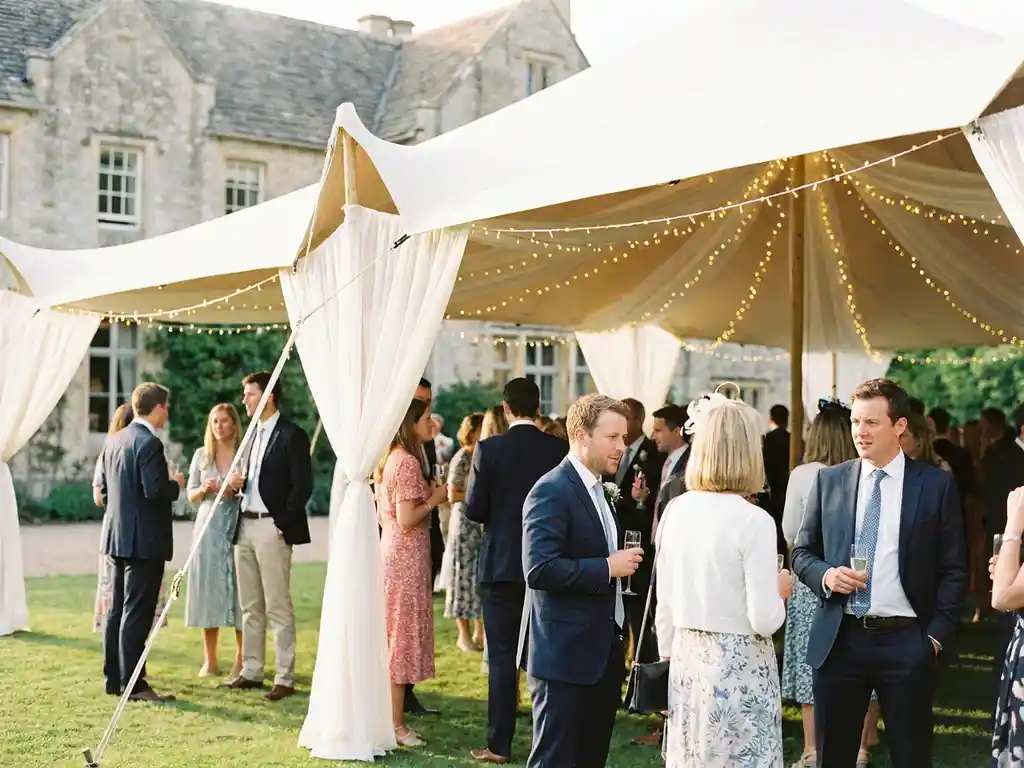 Witte stretch tent met sprookjesverlichting over bruiloftsgasten op groen gazon tijdens gouden uur.
