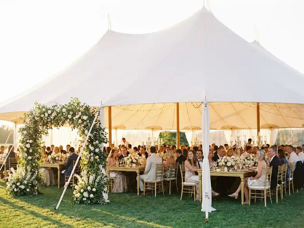 Elegante stretch tent met witte stoffen luifel boven een bruiloftsreceptie met gouden stoelen, bloemtafels en sfeervolle lichtjes in gouden avondlicht.