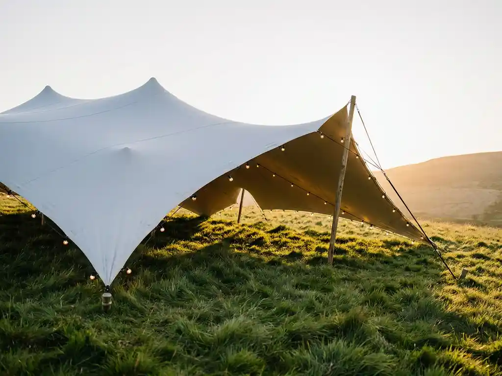 Witte stretchtent op glooiend grasveld met slingerverlichting in warm middaglicht, omgeven door groene heuvels.